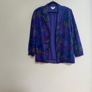 May include: A long-sleeve jacket featuring a vibrant purple, blue, and dark gray abstract floral pattern. The garment has a solid purple inner lining, a notched collar, and elasticized cuffs and hem. A white label with the text DEE is visible.