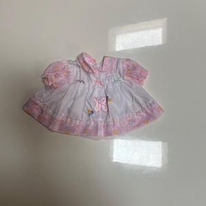 May include: A white doll dress with short puff sleeves, a Peter Pan collar, and a pink butterfly embroidery on the front. The dress features a floral patterned hem in shades of pink, yellow, and blue, with delicate lace trim and small ribbon bows.