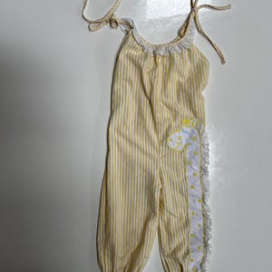 May include: Yellow and white striped baby jumpsuit with a giraffe applique. The jumpsuit features a ruffled neckline and trim, with tie straps. The giraffe applique is on the front, with a white and yellow design.