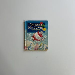 May include: A vintage children's book titled "Up Goes Mr. Downs" by Jerry Smath. The cover features a cartoon illustration of a character floating above a city with balloons. The book has a blue cover with red, yellow, and green balloons.