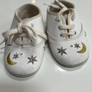 May include: A pair of white baby shoes with cream-colored laces. Each shoe features a gold crescent moon and star design. The shoes have a soft, padded interior and are designed for infants.