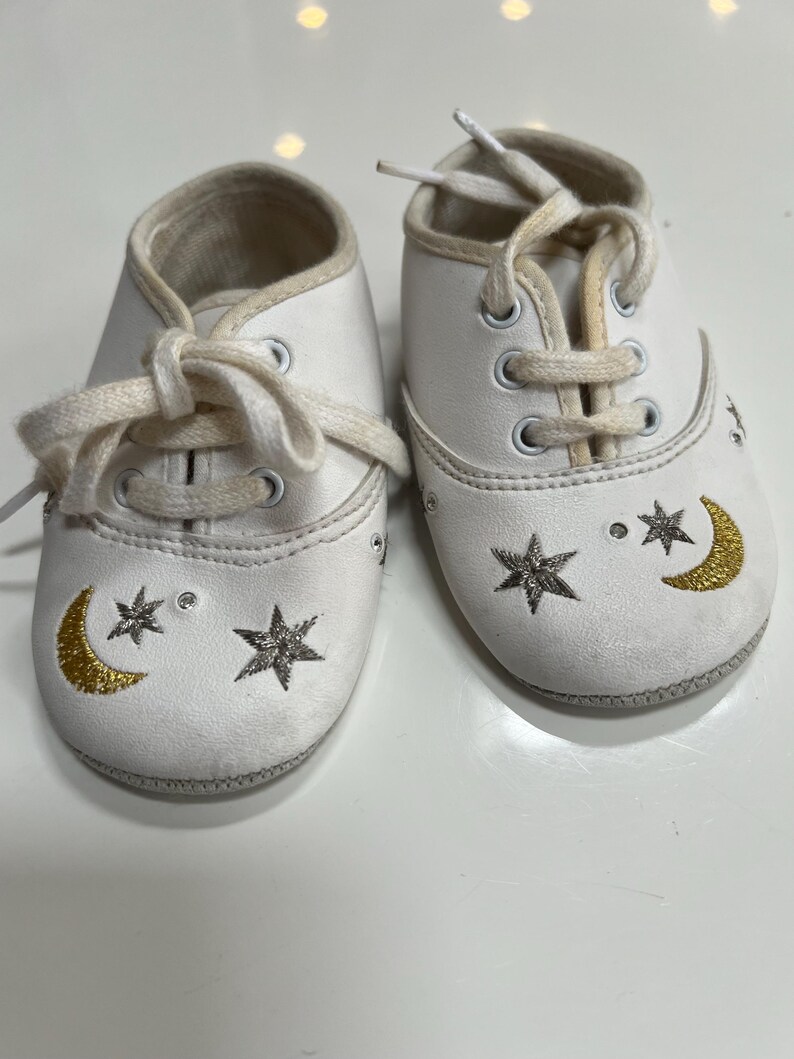 May include: A pair of white baby shoes with cream-colored laces. Each shoe features a gold crescent moon and star design. The shoes have a soft, padded interior and are designed for infants.