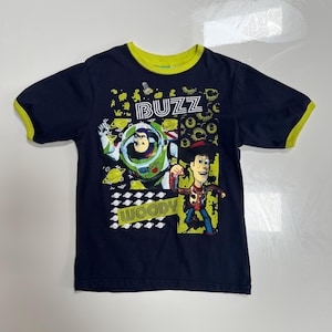 May include: Navy blue t-shirt featuring Buzz Lightyear and Woody from Toy Story. The shirt has a green collar and sleeve trim, with the word "BUZZ" and "WOODY" in a playful font.