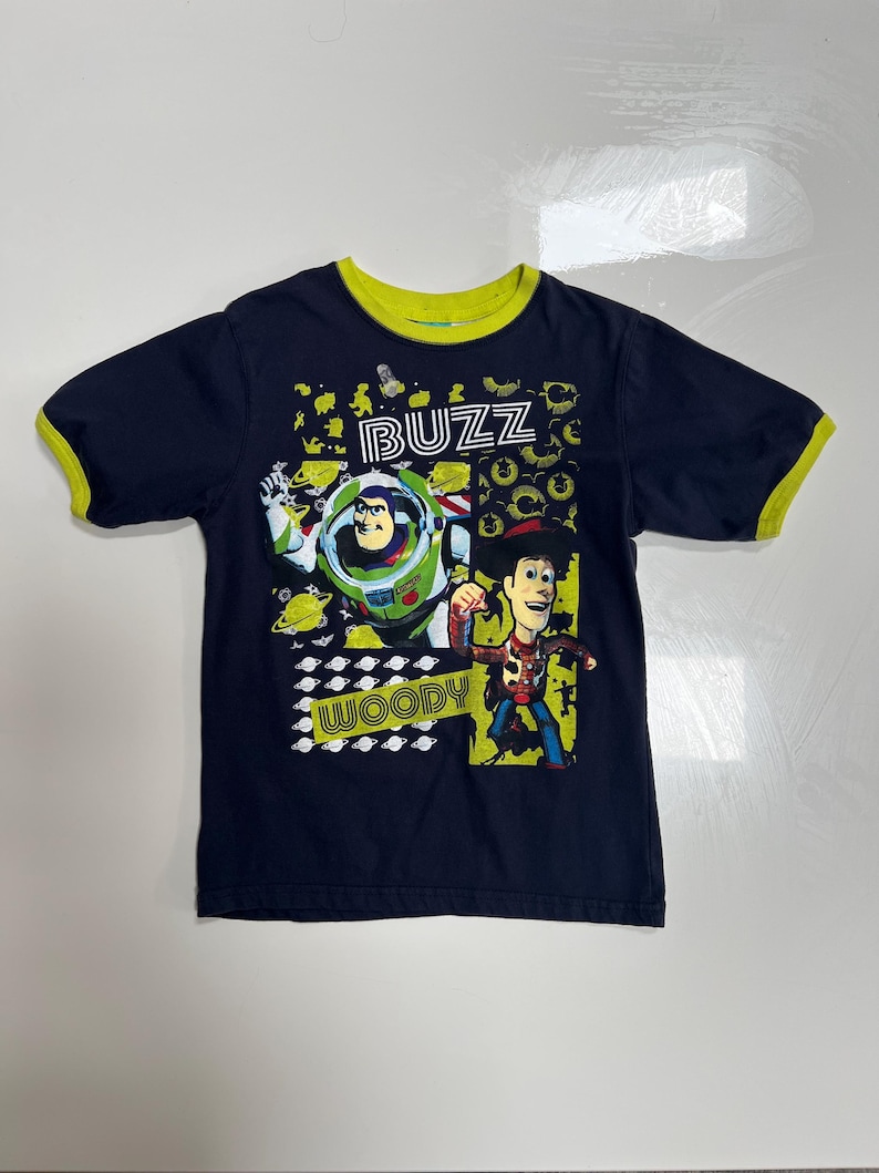 May include: Navy blue t-shirt featuring Buzz Lightyear and Woody from Toy Story. The shirt has a green collar and sleeve trim, with the word "BUZZ" and "WOODY" in a playful font.