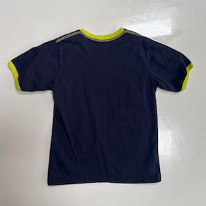 May include: A navy blue t-shirt with short sleeves, featuring a bright yellow trim around the collar and sleeve ends. The garment is likely made of cotton and is displayed flat on a white surface.