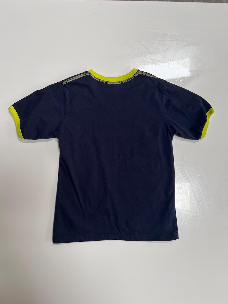 May include: A navy blue t-shirt with short sleeves, featuring a bright yellow trim around the collar and sleeve ends. The garment is likely made of cotton and is displayed flat on a white surface.