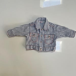 May include: A long-sleeved button-down shirt featuring a vertical blue, white, and black striped pattern with thin red accent stitching. The shirt includes a pointed collar, two chest pockets with button closures, and a front button-up placket.
