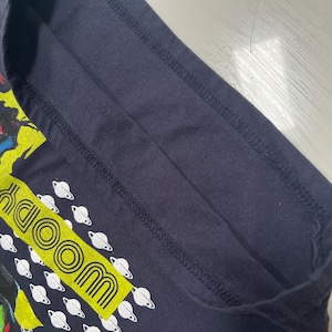 May include: Dark navy blue t-shirt with a cartoon graphic and the word "WOODY" in a yellow rectangle. White planet graphics are also present. The shirt's design is bright and colourful, perfect for fans of the character.