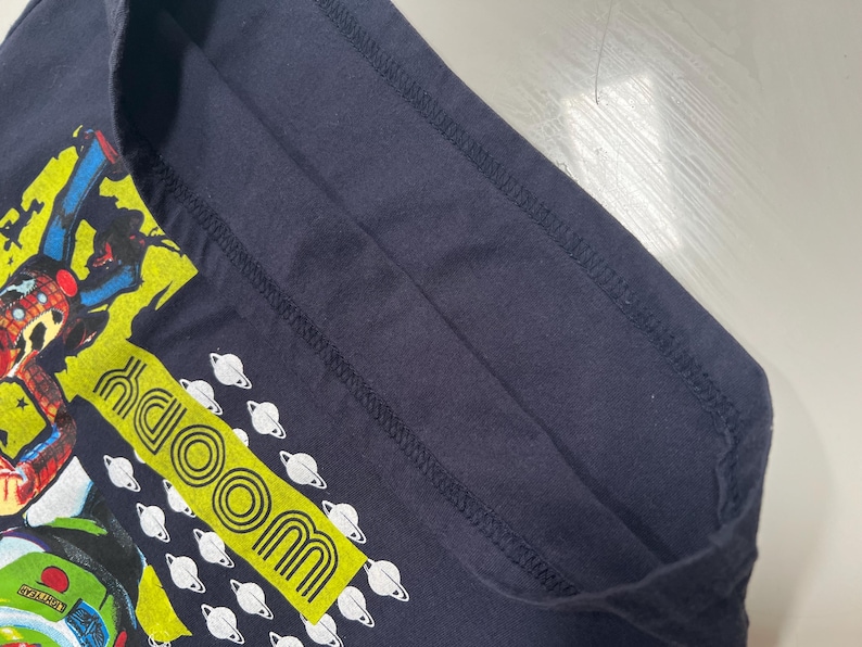 May include: Dark navy blue t-shirt with a cartoon graphic and the word "WOODY" in a yellow rectangle. White planet graphics are also present. The shirt's design is bright and colourful, perfect for fans of the character.