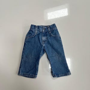 May include: Blue denim baby pants with a button closure, belt loops, and two front pockets. The pants have a classic five-pocket design and a straight leg. The denim is a medium wash with some fading.