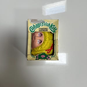 May include: A Cabbage Patch Kids costume and mask set in its original cardboard packaging. The box features a yellow plastic mask with blonde hair and a red bow, visible through a cutout. The packaging includes green text and graphics.