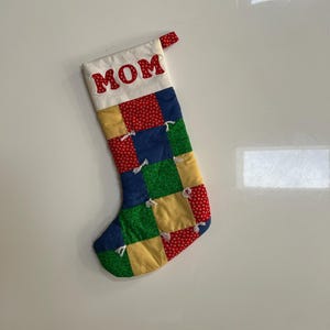 May include: A Christmas stocking with a patchwork design in red, blue, green, and yellow squares. The top of the stocking is white with the word "MOM" in red letters. A red loop is attached to the top.