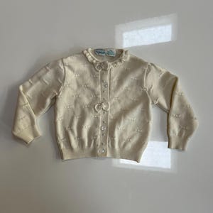 May include: Cream-colored cardigan with a ruffled collar and bow details. The button-down sweater features small bow-shaped embellishments and ribbed cuffs. The tag reads "Nolyn". A vintage-inspired piece.