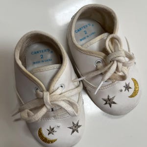 May include: A pair of white baby shoes with cream-colored laces. Each shoe features a gold crescent moon, silver stars, and a small rhinestone. The shoes are marked "Carter's" and size 2.