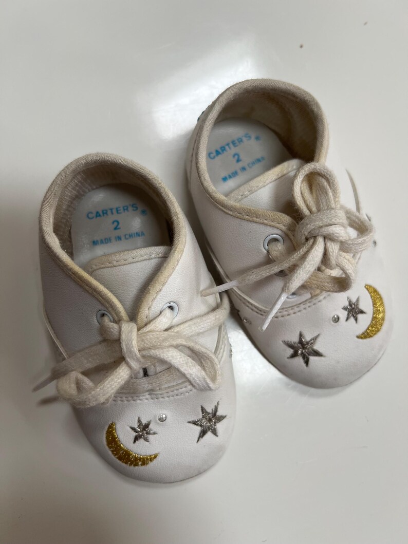 May include: A pair of white baby shoes with cream-colored laces. Each shoe features a gold crescent moon, silver stars, and a small rhinestone. The shoes are marked "Carter's" and size 2.