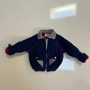 May include: A navy blue corduroy jacket with a gray collar and red accents. The jacket has a zipper closure, two front pockets, and red and gray plaid cuffs. The jacket has the text "Park City Kids" on the collar.