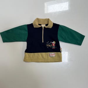 May include: A long-sleeve pullover with a tan collar and hem, dark blue center, and green sleeves. It features a half-zip closure, a graphic of a person blowing bubbles with the text "CATCH ME IF YOU CAN BUBBLES!", and a "KIDSPAY" label.