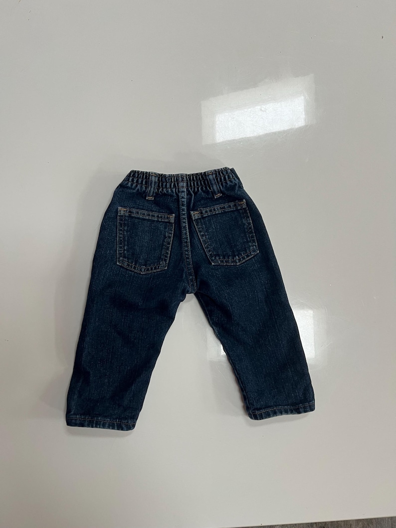 Op de afbeelding: Donkerblauwe spijkerbroek voor een peuter, met twee achterzakken en een elastische tailleband. De jeans is gemaakt van een stevig denim materiaal en heeft een klassieke, casual stijl.