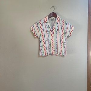 May include: A short-sleeved button-down shirt in white featuring a repeating vertical pattern of red zig-zag lines, blue wavy lines, and green dots. The shirt has a classic collar and a label at the inner neckline that reads FASE.