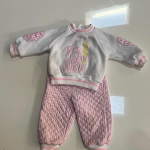 May include: A white and pink children's pajama set. The top has a pink teddy bear graphic and pink elbow patches. The pants are quilted pink with a diamond pattern. The set has pink and white striped trim.
