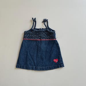 May include: A blue denim dress with a dark blue crocheted bodice and spaghetti straps. A pink band accents the waist, and a red heart applique is on the skirt. The dress is designed for a child.