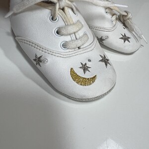 May include: White baby shoes with cream-colored laces. The shoes are decorated with gold glitter crescent moons and star designs. The shoes have a classic sneaker style with a rounded toe.