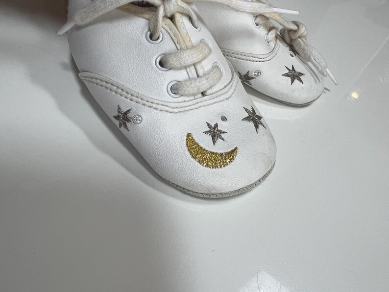 May include: White baby shoes with cream-colored laces. The shoes are decorated with gold glitter crescent moons and star designs. The shoes have a classic sneaker style with a rounded toe.