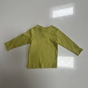 May include: A light green long-sleeved shirt with a mock neck. The shirt has small, scattered embellishments across the front. The garment is made of a soft material and is suitable for children's wear.
