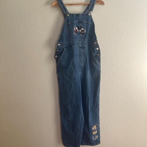 May include: Blue denim overalls with Mickey Mouse and Minnie Mouse embroidered on the bib pocket. The overalls have adjustable straps, side buttons, and pockets. The bottom of the left leg has embroidered character patches. The overalls are hanging on a wooden hanger.