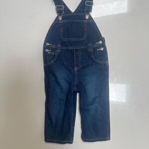 May include: Dark blue denim overalls with adjustable straps and metal hardware. The bib overalls have two front pockets and red stitching details. The garment is designed for children.