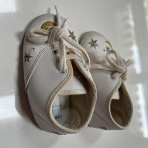 May include: A pair of white baby shoes with star and moon designs. The shoes have white laces and a gold glitter accent on the top. The shoes are made of a soft material and are designed for infants.