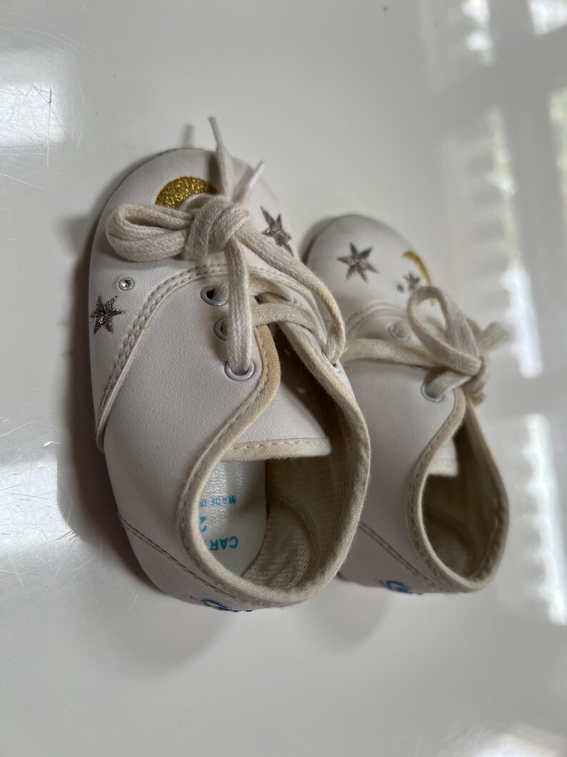 May include: A pair of white baby shoes with star and moon designs. The shoes have white laces and a gold glitter accent on the top. The shoes are made of a soft material and are designed for infants.