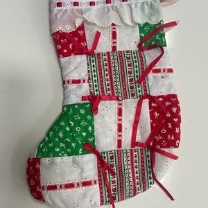 May include: A handmade Christmas stocking with a patchwork design. The stocking features red, green, and white fabric with floral and geometric patterns. Red ribbons are tied on the front. The top has a white lace trim.