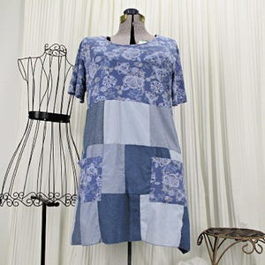 May include: A blue patchwork dress with short sleeves and two pockets. The top portion features a floral pattern, while the lower section is a mix of solid and patterned blue fabrics. The dress is displayed on a dress form.
