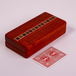 May include: A wooden cribbage board with a rich, reddish-brown finish. The board features inlaid decorative stripes and is paired with a standard playing card. The game board is rectangular and appears to be closed, suggesting it is a storage box.