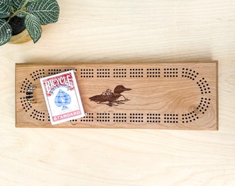 May include: A wooden cribbage board with a loon design and a deck of Bicycle playing cards. The board has a dark wood finish and a light wood border. The playing cards are red and blue.