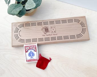 May include: A wooden cribbage board with a black and white design. The board has a cutout for cards and the text "The Perfect Hand" is engraved on the board. A deck of Bicycle playing cards and a red velvet pouch are also pictured.