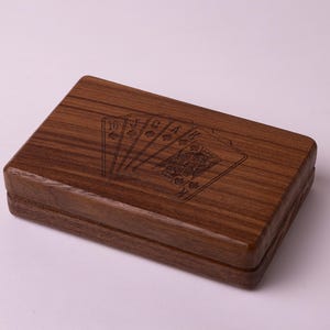 May include: A rectangular wooden playing card box with a dark brown finish. The top of the box features an engraved design of playing cards, including the ten of spades, queen of diamonds, and ace of hearts. The box is closed.