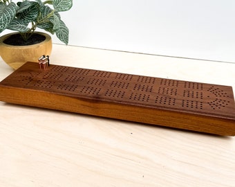 May include: A wooden cribbage board with a brown stain. The board has a grid of holes for pegs and is ready for a game.