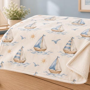 May include: A cream-colored blanket featuring a repeating pattern of sailboats with blue and white sails, light blue waves, small yellow suns, and blue birds. The blanket has a finished edge and is folded to show the soft texture.