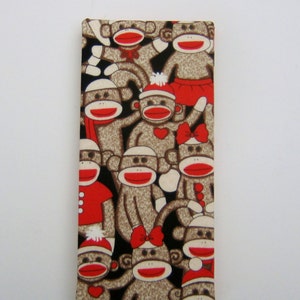 May include: A rectangular fabric item featuring a repeating pattern of sock monkeys. The monkeys have brown bodies, red mouths, and white faces. Some monkeys wear red hats, scarves, or bows. The background is black.