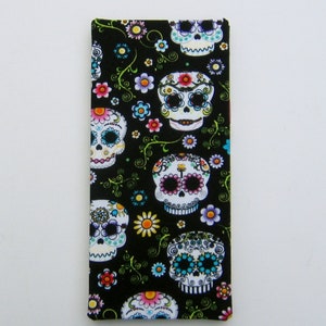 May include: A black rectangular fabric item featuring a repeating pattern of sugar skull designs. The skulls are white with colorful floral and decorative accents. Green vines and colorful flowers are scattered across the background.
