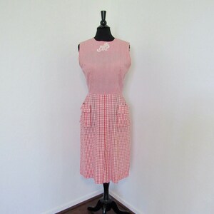 Pode incluir: Um vestido sem mangas de xadrez vermelho e branco com uma flor branca bordada na gola. O vestido tem bolsos nas laterais e é exibido em um manequim preto.
