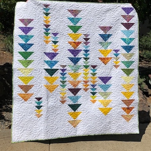 May include: A white quilt with a repeating pattern of colorful triangles in shades of purple, blue, green, yellow, and orange. The quilt has a green border and a detailed, swirling stitch pattern across the white background.