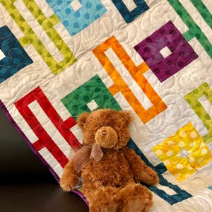 May include: A colorful patchwork quilt with a geometric pattern of squares and lines. A brown teddy bear is sitting on the quilt.