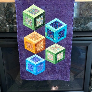 May include: A purple quilted wall hanging with five colorful fabric cubes arranged in a pyramid shape. The cubes are made of different patterned fabrics and have a 3D effect.