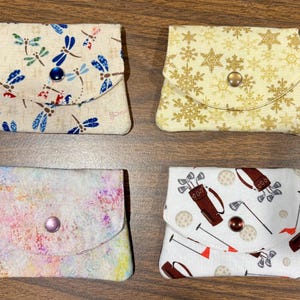 May include: Four small fabric pouches with snap closures. One features a dragonfly print on cream, one has gold snowflakes on yellow, one displays a multicolored abstract watercolor pattern, and one shows a golf-themed print on white fabric.