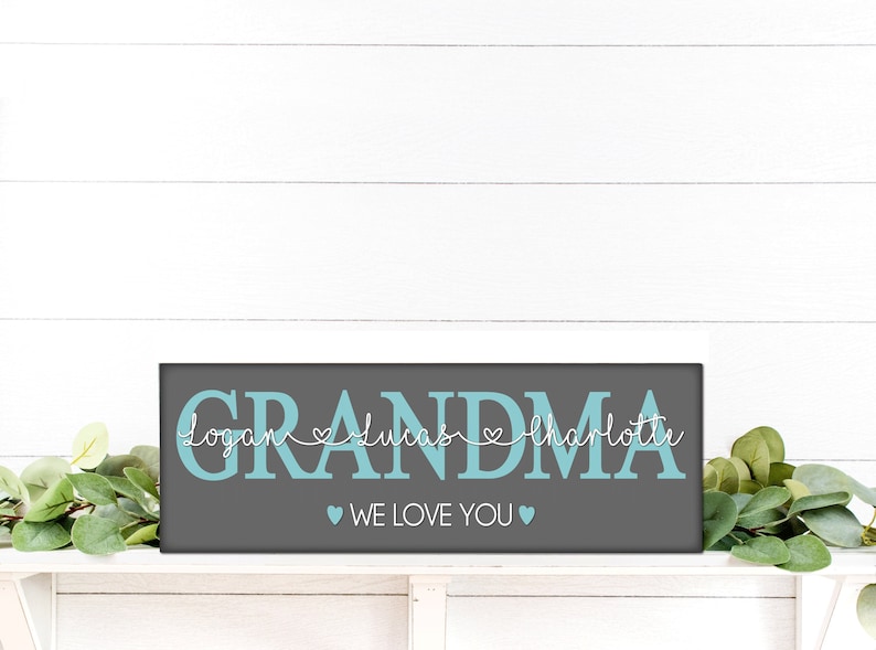 May include: A rectangular wooden sign with the word "GRANDMA" in teal block letters. Below the word are the names "Logan", "Lucas", and "Charlotte" in white script. The phrase "WE LOVE YOU" is below the names.