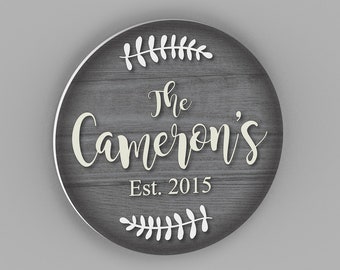 May include: A round wooden sign with a gray woodgrain background and white lettering. The sign reads "The Cameron's Est. 2015" with white leafy sprigs above and below the text.
