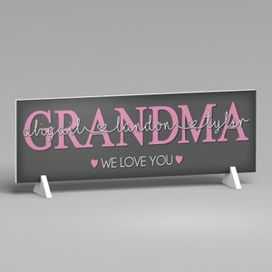 May include: A rectangular sign with the word "GRANDMA" in pink letters on a dark gray background. The names "abigail landon tyler" are above the word "GRANDMA". The phrase "WE LOVE YOU" is below the word "GRANDMA". The sign is supported by two white stands.